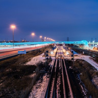 View of the Pickering GO Pedestrian Bridge at dusk. Programmable LED lighting animates and accentuates the curved form of the metal scrim – giving it movement and shimmer in the night sky. Photo by Michael Muraz.